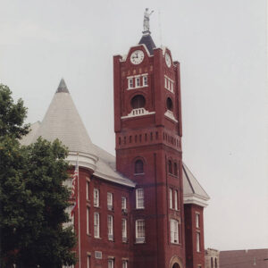 Multistory red brick building with large clock tower with statue atop it and a round tower next to it
