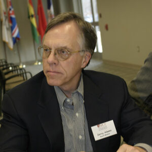 White man in suit and tie and glasses sitting at table and holding a pen