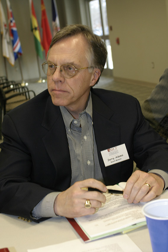 White man in suit and tie and glasses sitting at table and holding a pen