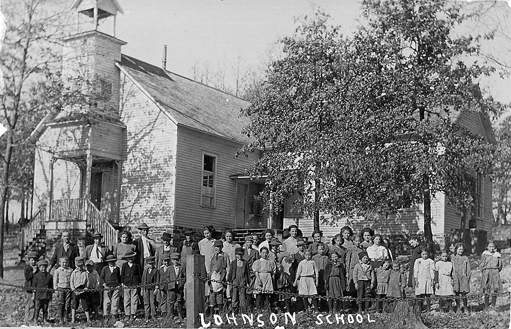Large group of white children and white adults standing in front of multistory wooden building with bell tower