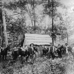 Group of white men and horses gathered in front of small wooden building