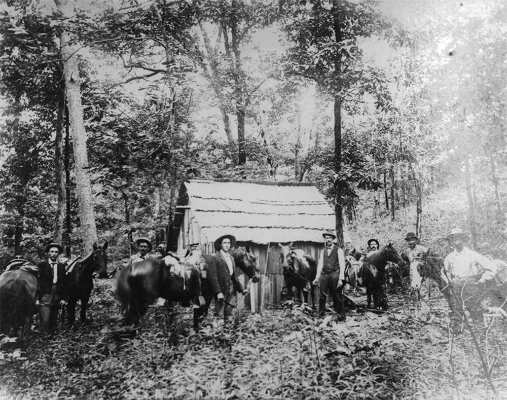 Group of white men and horses gathered in front of small wooden building