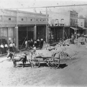 Row of storefront buildings with white men standing in front and horse-drawn wagons in the street