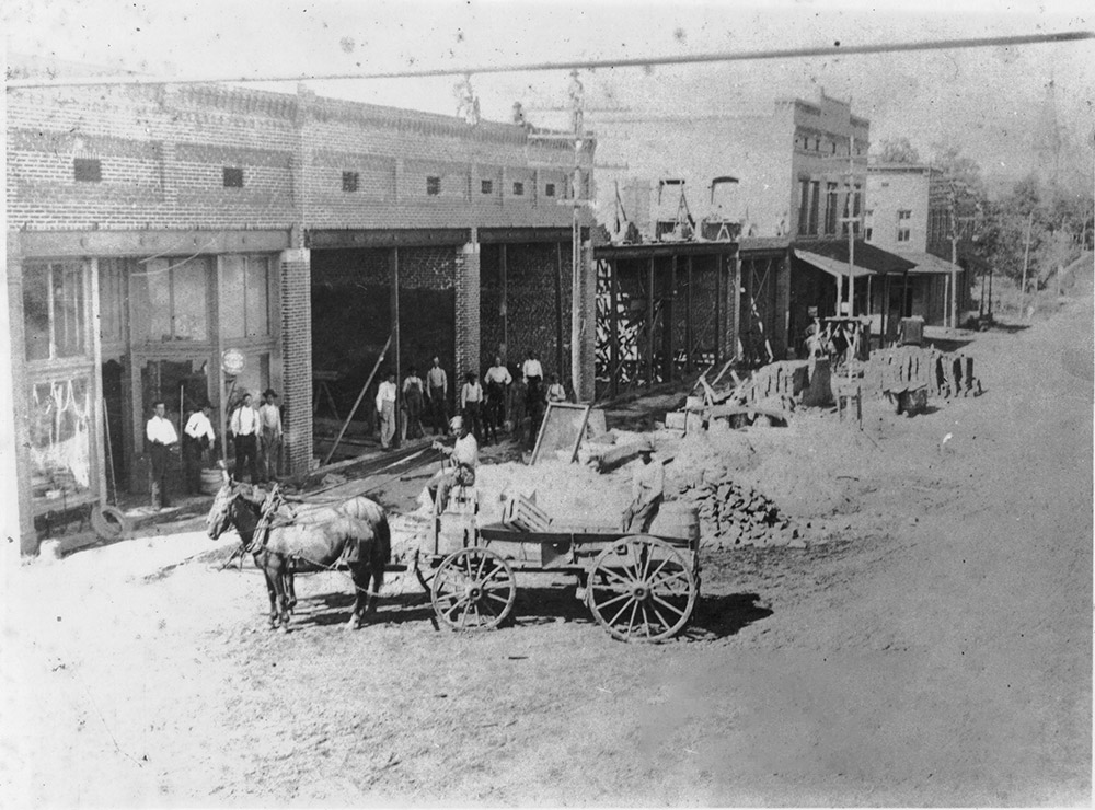 Row of storefront buildings with white men standing in front and horse-drawn wagons in the street