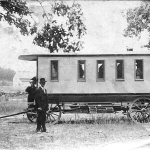 White people standing around horse-drawn coach with steps protruding from the back with priest standing at top