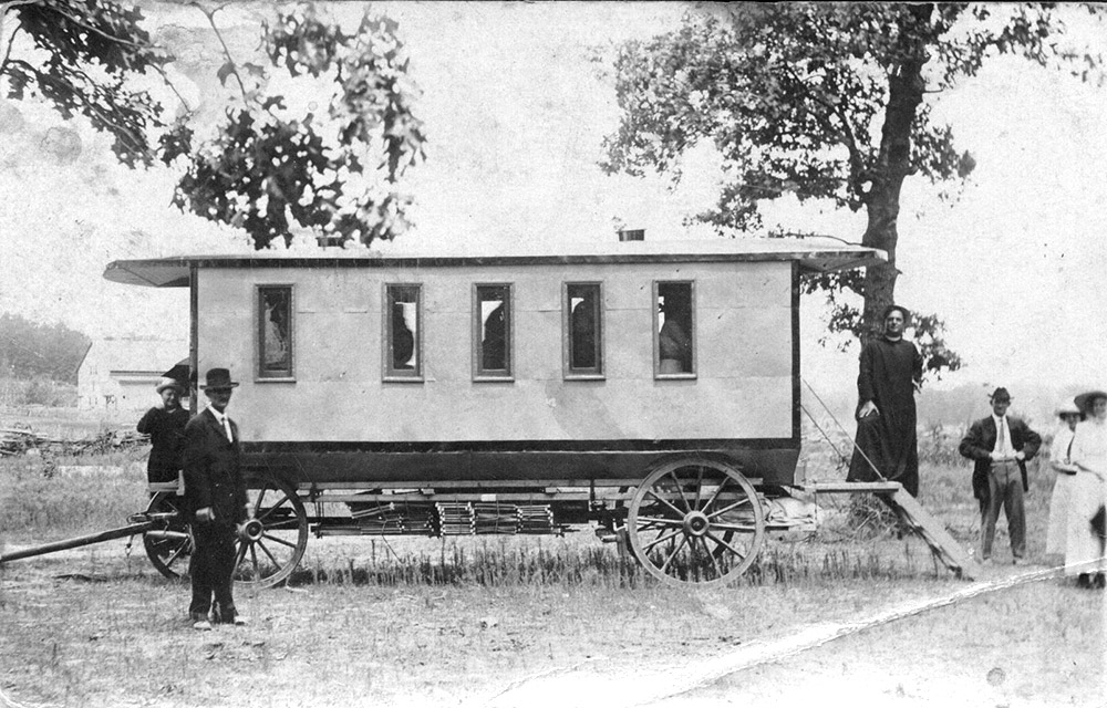White people standing around horse-drawn coach with steps protruding from the back with priest standing at top