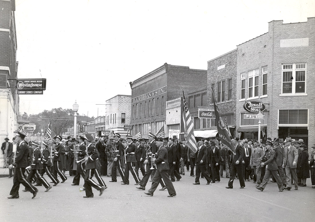 Uniformed white boys and men some with instruments and flags parading down street lined with multistory storefront buildings