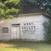 Single story abandoned white wooden building with "West Valley Community Building" printed on it in black letters