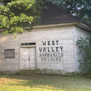 Single story abandoned white wooden building with "West Valley Community Building" printed on it in black letters