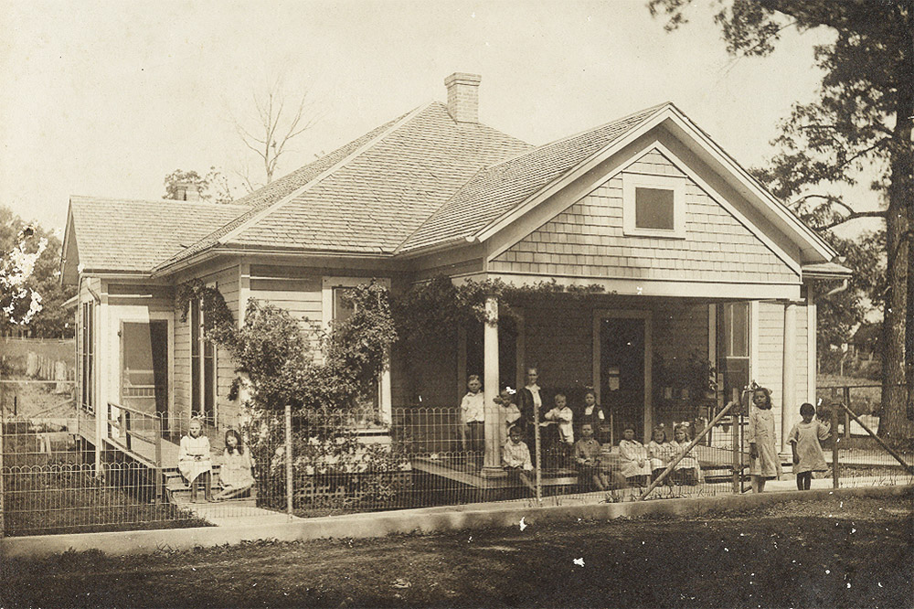 Single story white wooden house with more than a dozen white children sitting and standing