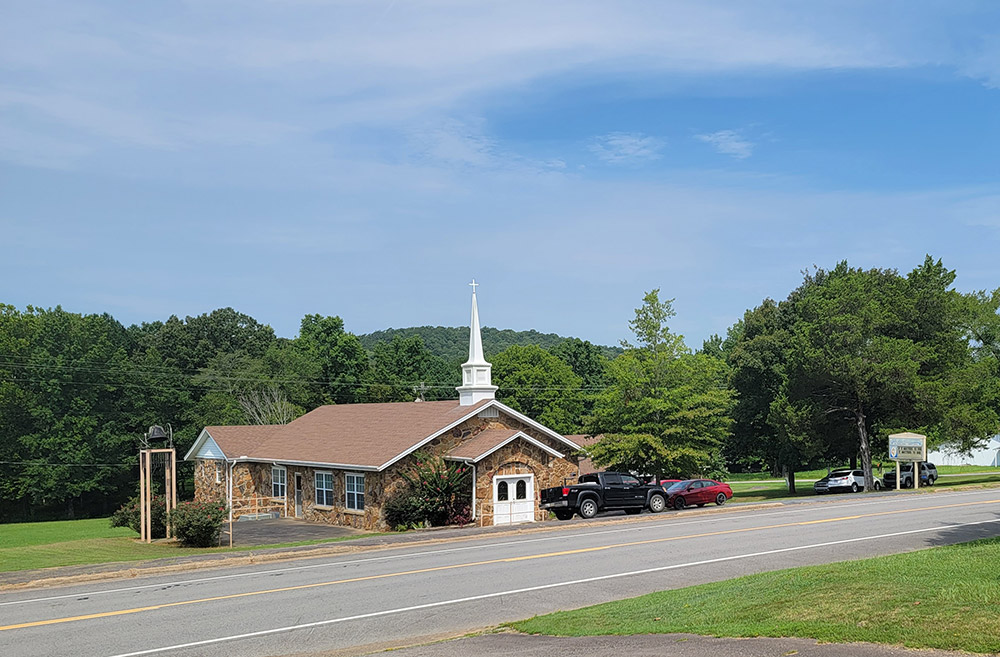 Rock church building with steeple situated by road with trees in background