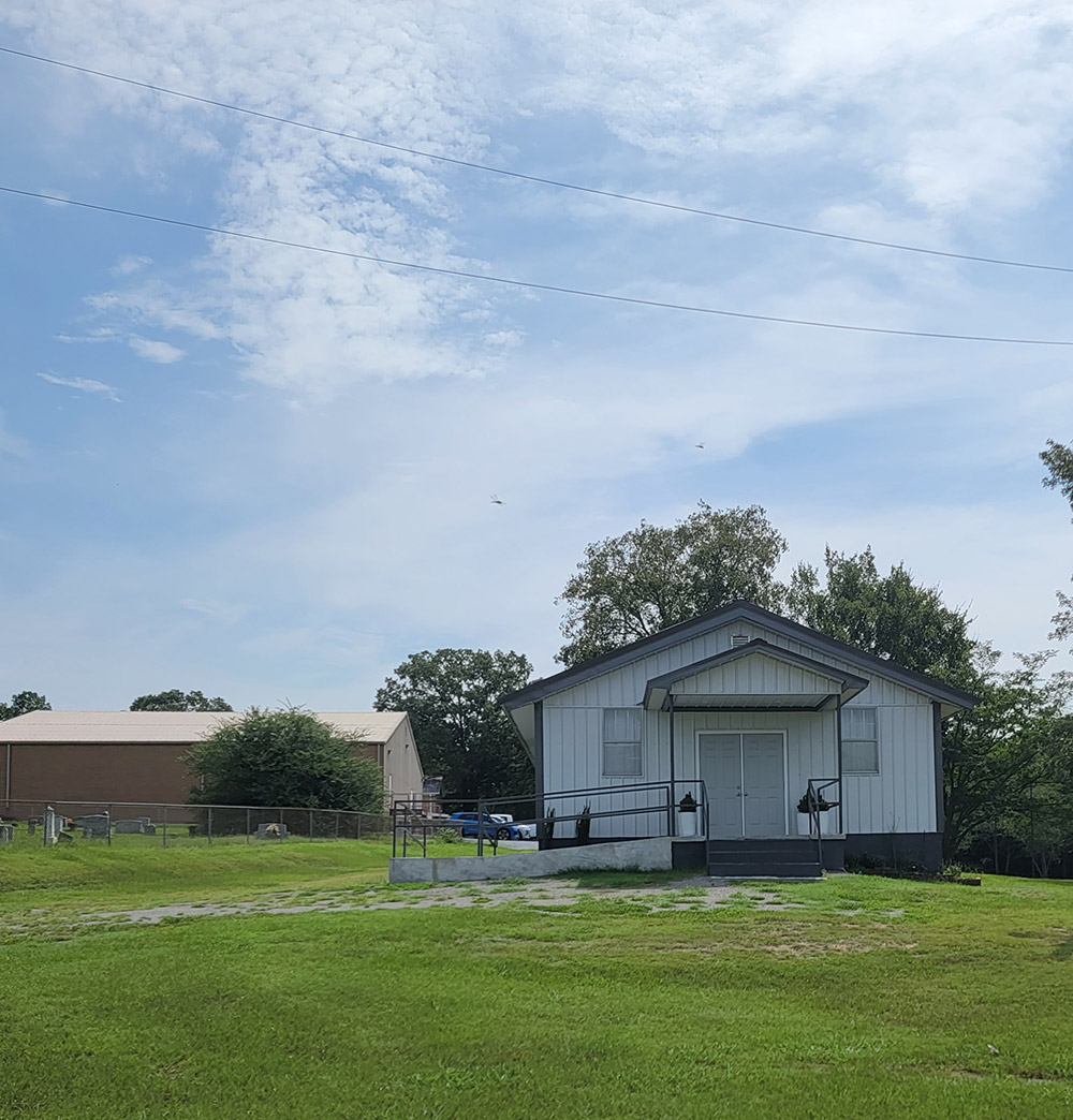 Single story white metal building with access ramp and grass in front and cemetery beside