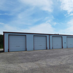 White metal building with four garage doors and one entrance door with gravel lot
