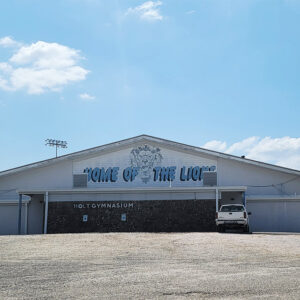 White gymnasium building with "Home of the Lions" painted on it and parking lot
