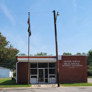 Single story red brick post office building with American flag on pole and parking lot