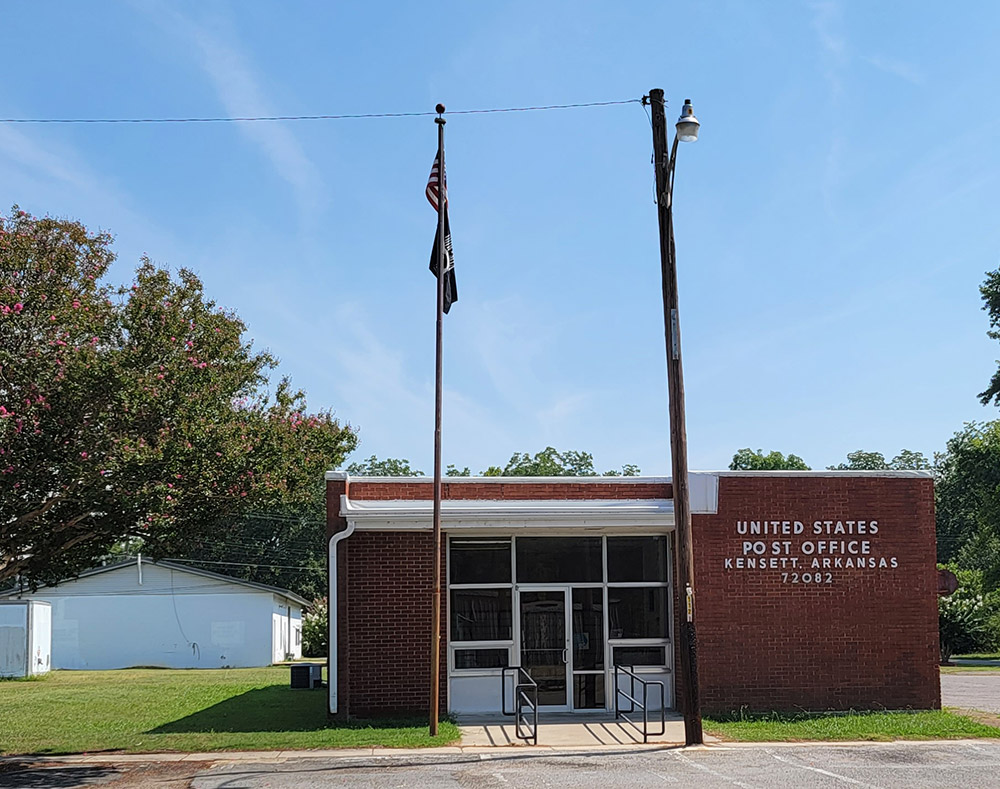 Single story red brick post office building with American flag on pole and parking lot