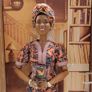 Doll of African American woman in black and orange flowery dress and head covering holding a book in front of her