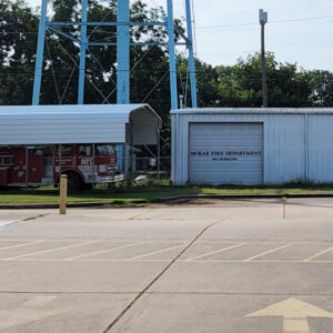 Fire engine beneath metal shelter next to single story white metal building with garage door
