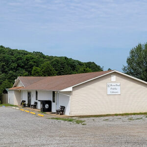Single story tan wooden building with sign saying "Rose Bud Public Library" and gravel parking lot