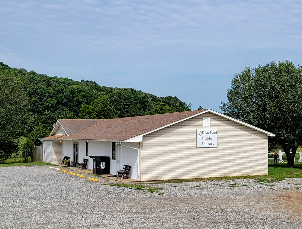 Single story tan wooden building with sign saying "Rose Bud Public Library" and gravel parking lot
