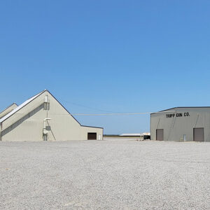 Enormous tan metal buildings with "Tripp Gin Company" printed on one amid immense gravel-covered stretch of land