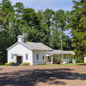 Single story white wooden church building with front and side entrances and parking lot