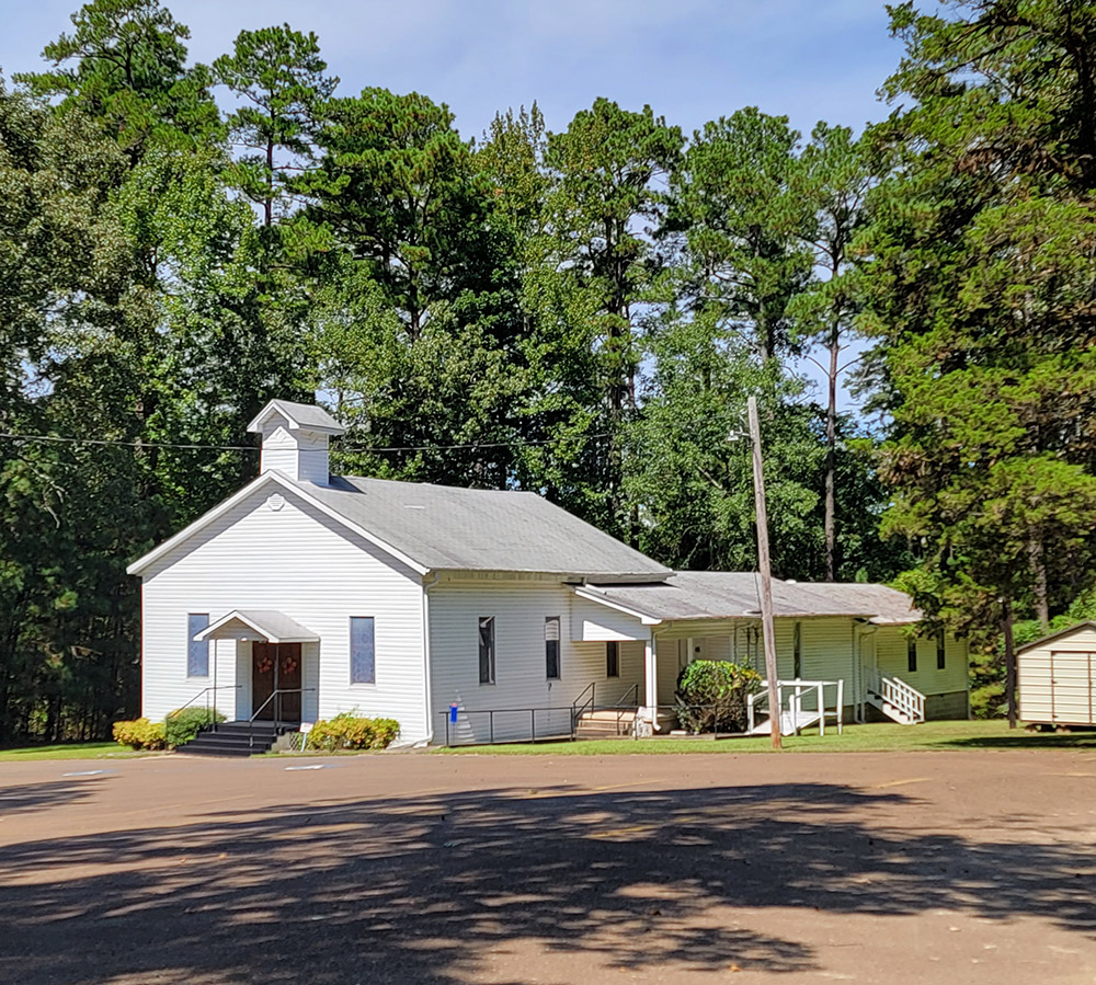 Single story white wooden church building with front and side entrances and parking lot