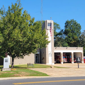 Multistory tan brick building with fire trucks in garages and monument in front