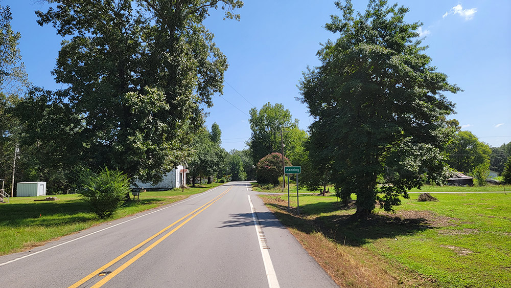 Rural road with scattered trees and houses and sign saying "Manning"