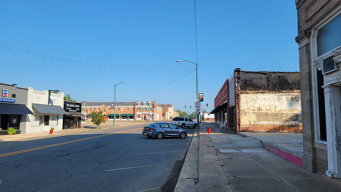 Single story storefront buildings along road with multistory orange brick building in distance