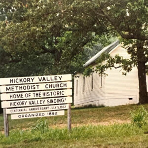 Single story white wooden church building and sign saying "Home of the Historic Valley Singing"