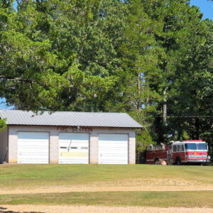 Single story white concrete block building with three garage bay doors "Fire Station" and fire truck parked outside