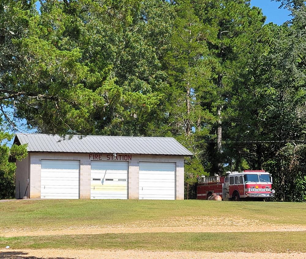 Single story white concrete block building with three garage bay doors "Fire Station" and fire truck parked outside