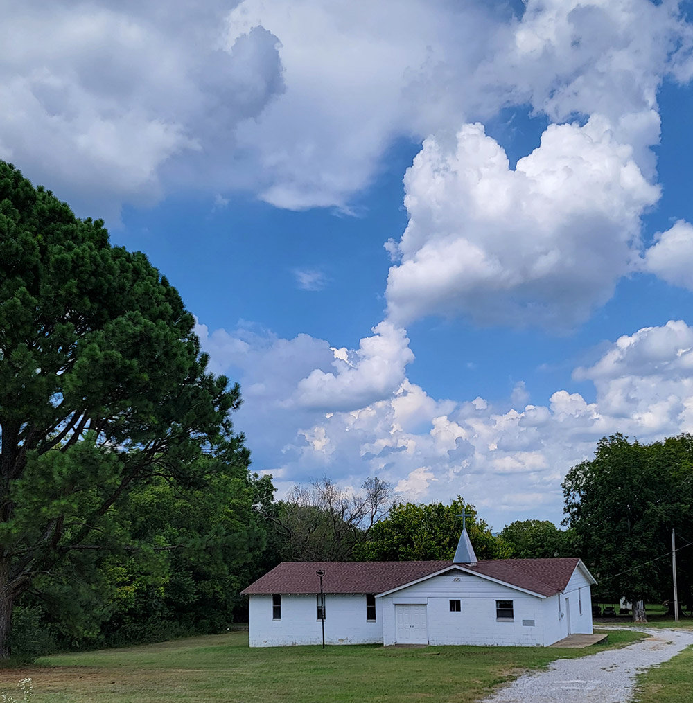 Single story white wooden church building with steeple and gravel road and trees in background