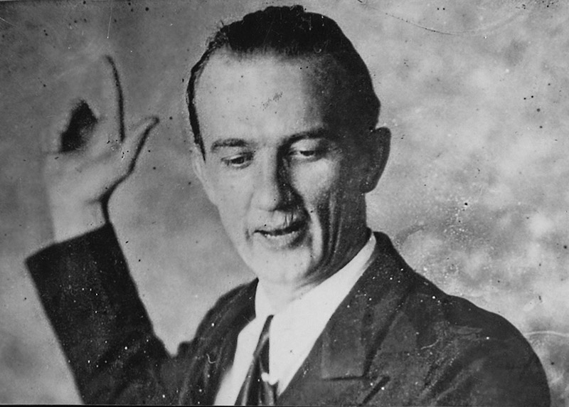 White man in suit and tie looking to the side with his arm and finger raised