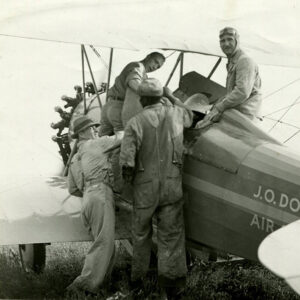 Men performing tasks around an airplane with one man in the cockpit with goggles on his head