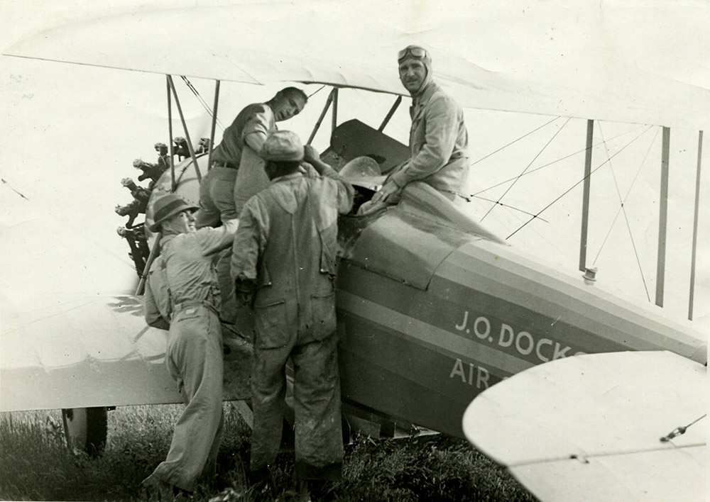 Men performing tasks around an airplane with one man in the cockpit with goggles on his head
