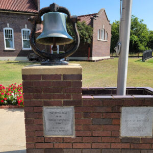 Brass bell on a brick stand next to another brick stand holding a flagpole in front of a brick building