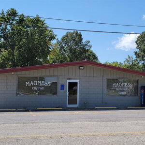 Single story gray concrete block building with sign saying "Magness city hall" with parking lot