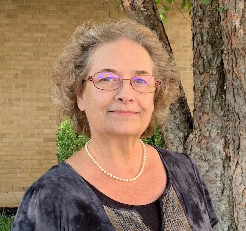 White woman wearing glasses and pearls standing in front of a tree and a brick wall