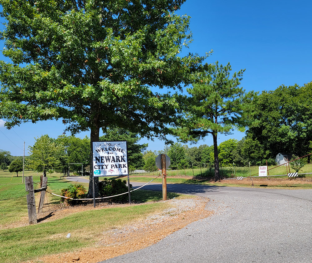 Road leading to park with grass and trees on both sides and sign saying "Welcome to Newark City Park"