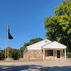 Single story tan brick post office building with flags on flagpole and parking lot