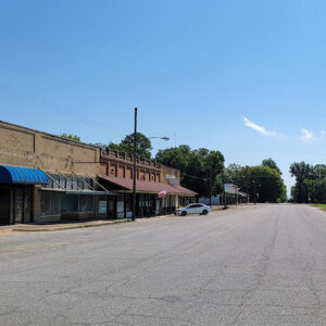 Street with mostly abandoned storefront buildings on one side and railroad tracks next to agricultural buildings on the other side
