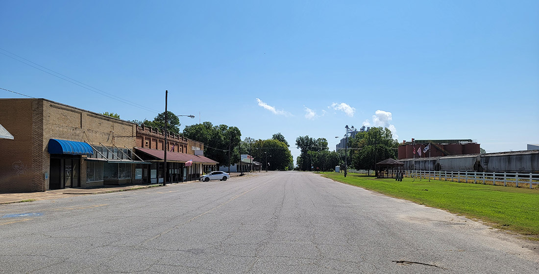 Street with mostly abandoned storefront buildings on one side and railroad tracks next to agricultural buildings on the other side