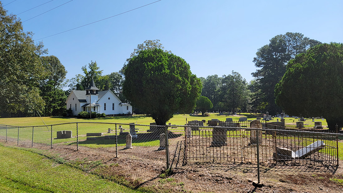 White country church building with bell tower in distance with cemetery and trees in foreground