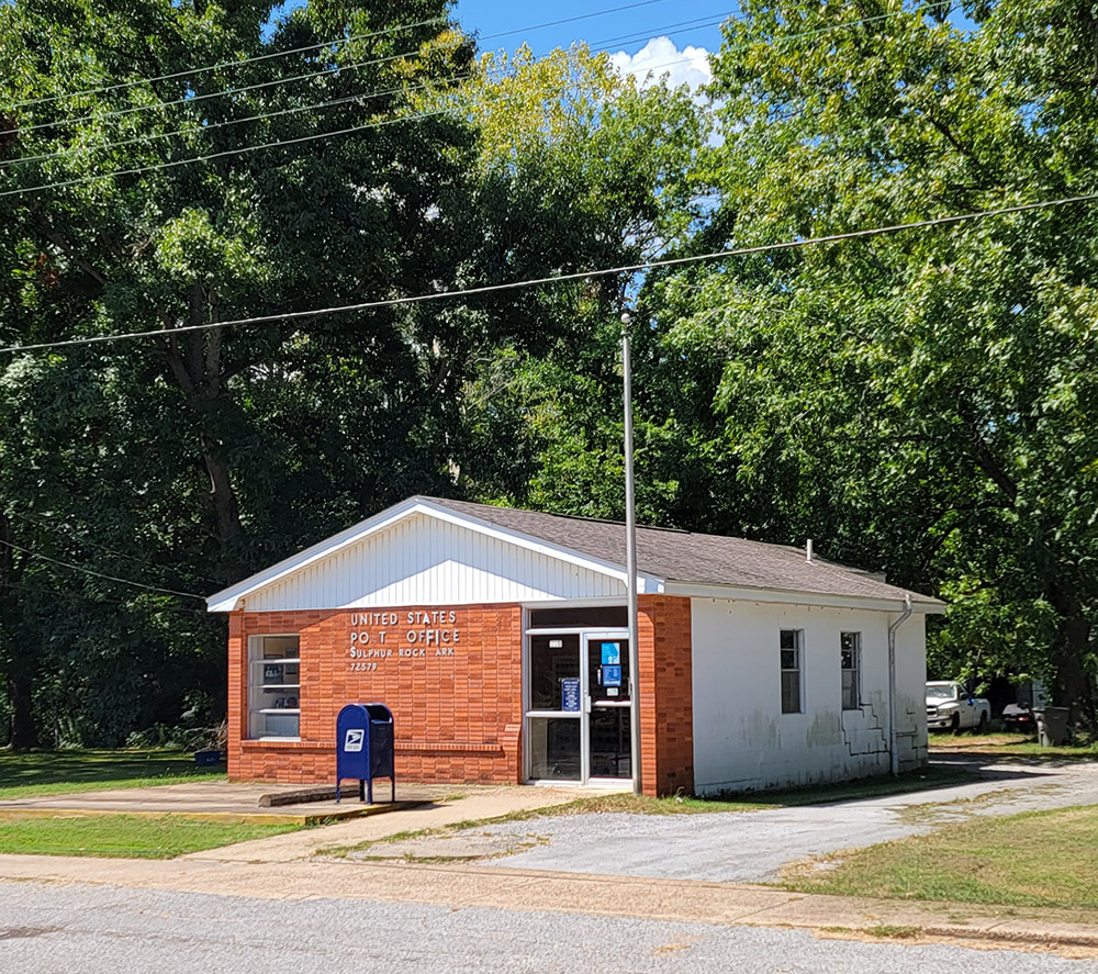 Single story post office building with red brick front and trees in background