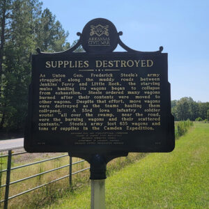 Metal sign saying "Supplies Destroyed" and giving information with fence and road in background
