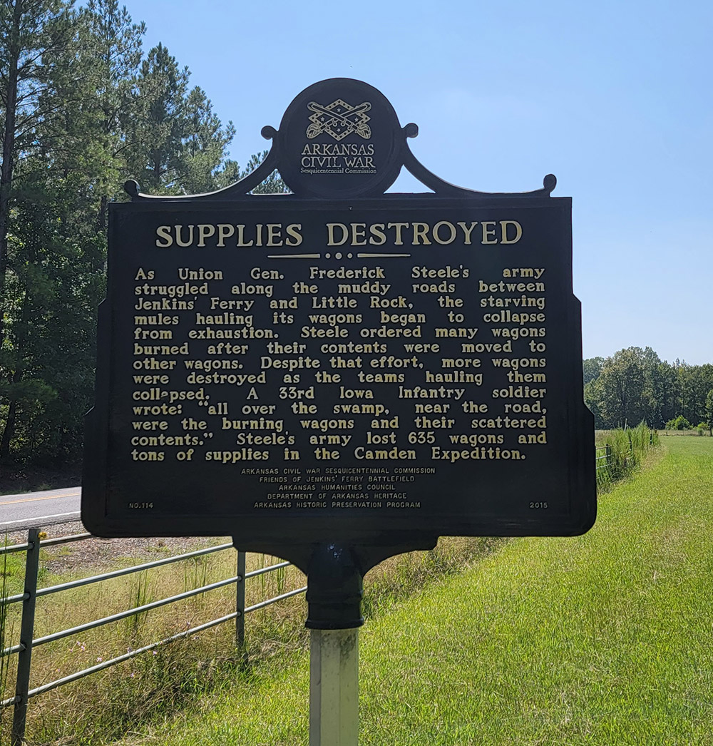 Metal sign saying "Supplies Destroyed" and giving information with fence and road in background