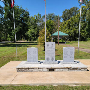 Three stone slabs with writing standing vertical and flagpoles in park setting with trees in background
