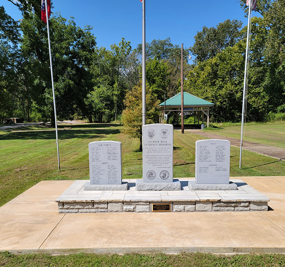 Three stone slabs with writing standing vertical and flagpoles in park setting with trees in background